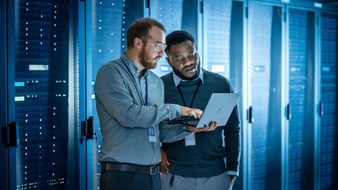 IT pros use laptop in data center and work next to server racks, running diagnostics/doing maintenance